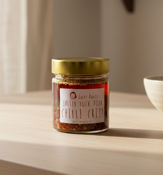 A glass jar of Just Kasz Salted Duck Yolk Chilli Crisp with a gold lid and white label, sitting on a dark countertop with a blurred kitchen background.