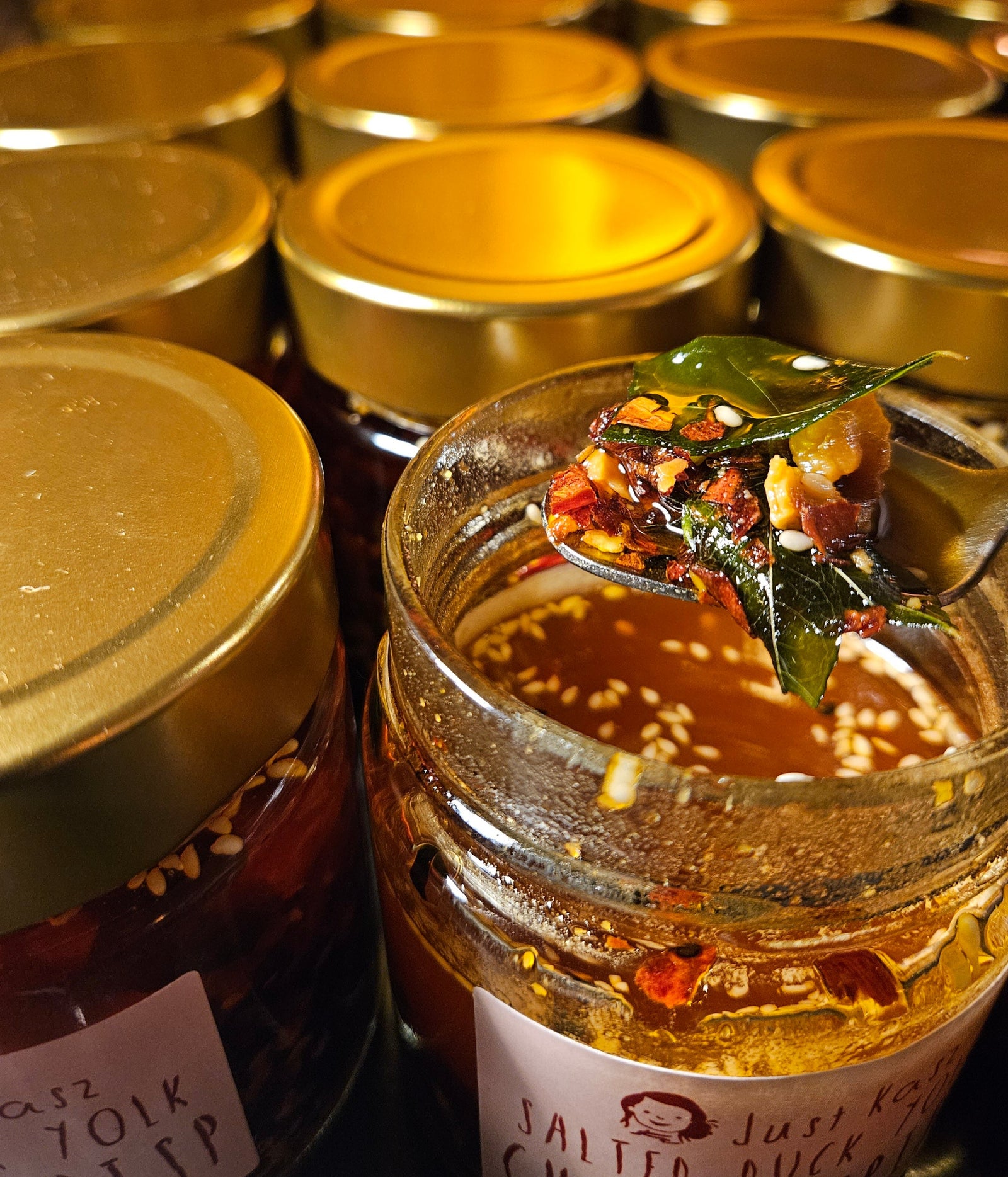 A close-up of a glass jar of Just Kasz Salted Duck Chilli Crisp with a spoonful of the spicy red oil and ingredients, surrounded by other jars with gold lids.