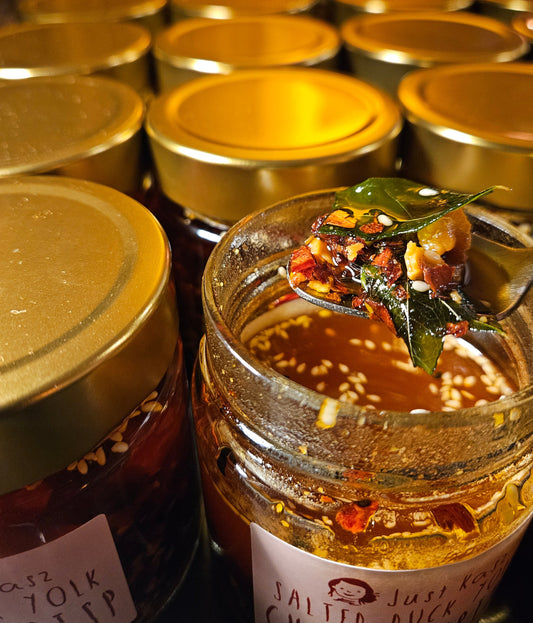 A close-up of a glass jar of Just Kasz Salted Duck Chilli Crisp with a spoonful of the spicy red oil and ingredients, surrounded by other jars with gold lids.